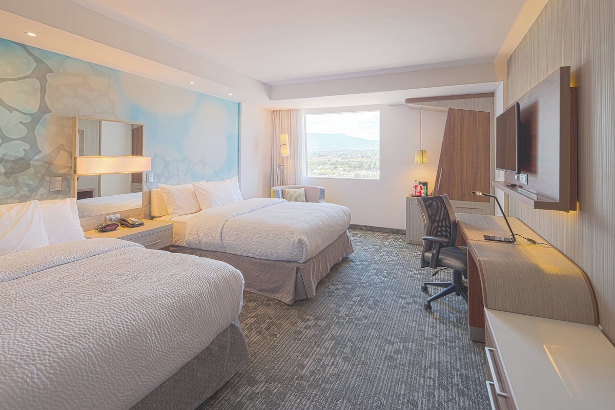 A hotel room with two beds, a desk with a chair, a TV, and a large window offering a city view.