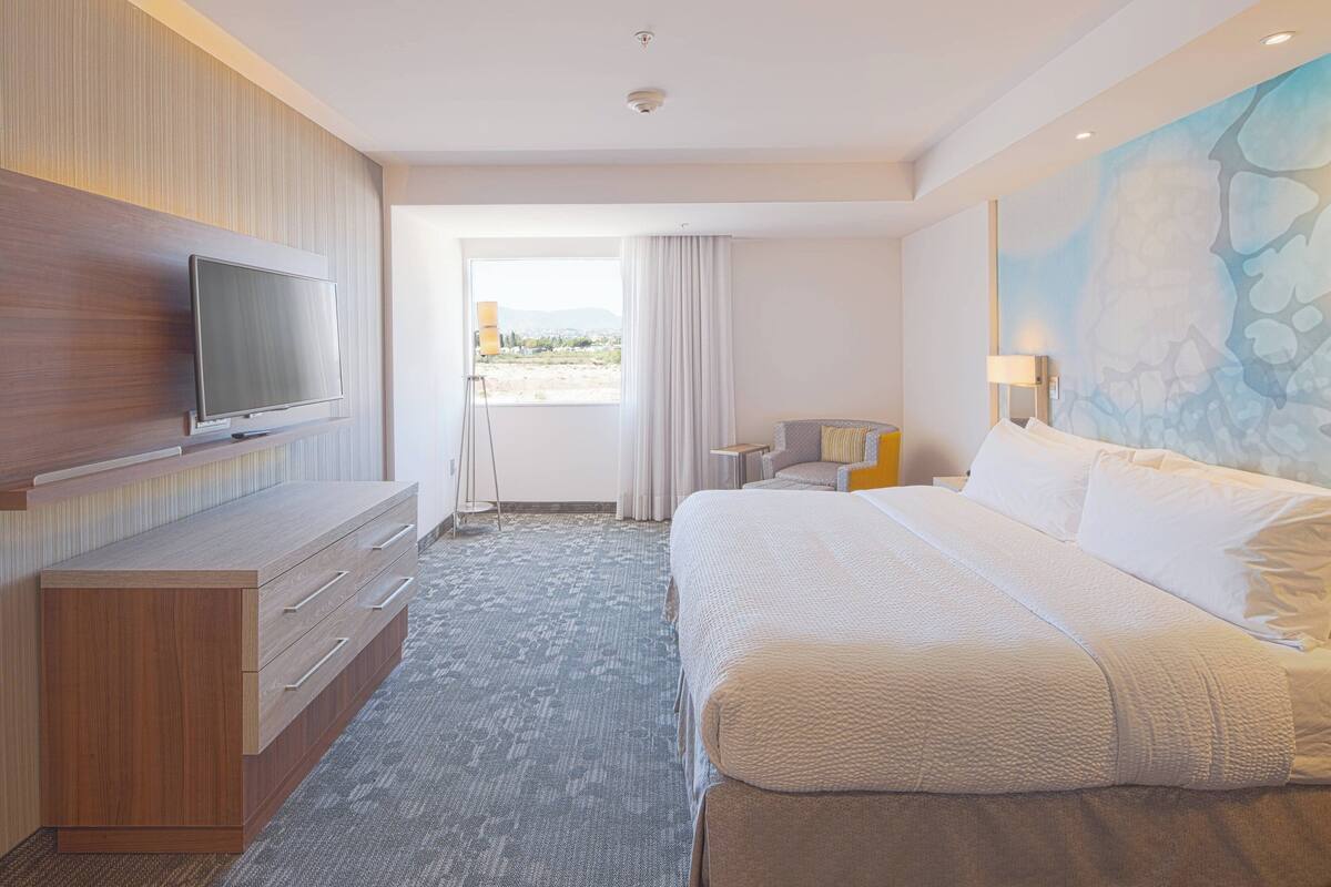 A hotel room with a large bed, a flat-screen TV mounted on a wooden cabinet, a small desk with a lamp, and a view of the outdoors through a door.