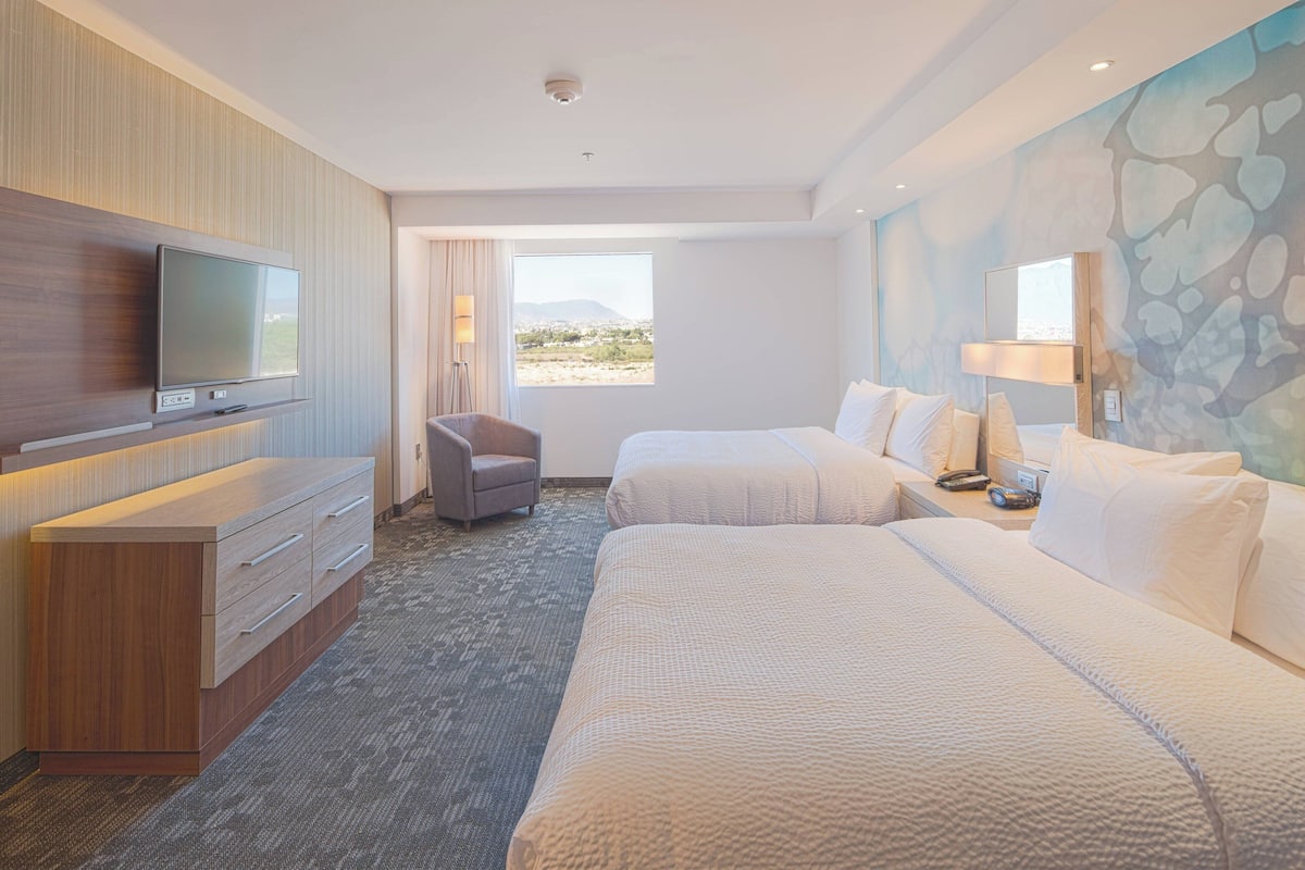 A hotel room with a flat-screen TV mounted on the wall, a desk with a lamp, two beds, a chair, and a large window offering a view of the desert.