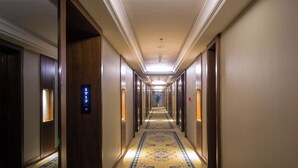 Un largo y estrecho pasillo de hotel con puertas de madera a ambos lados, una alfombra estampada y una lámpara en el techo.