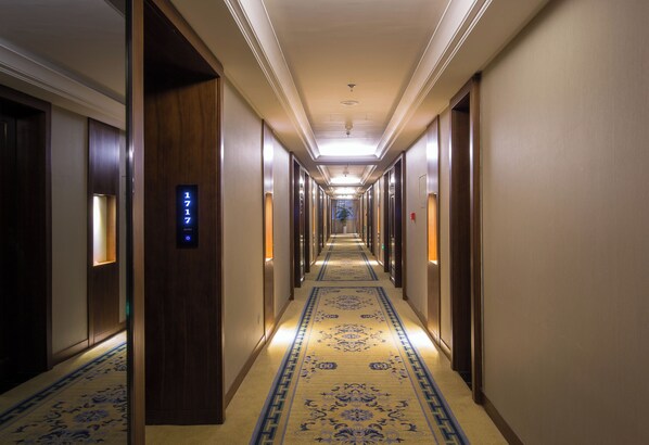 Un long couloir étroit d’hôtel, avec des portes en bois de chaque côté, une moquette à motifs et un luminaire au plafond.