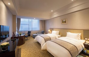 Habitación de hotel con dos camas, un escritorio, una silla, un televisor y un ventanal grande con cortinas.