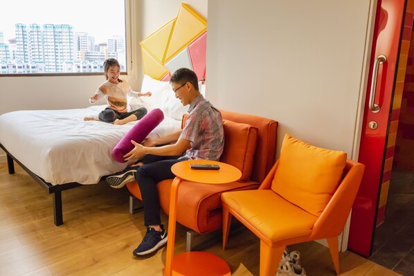 A modern hotel room with a bed, orange seating, a small table, and a city view.