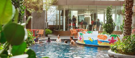 An indoor swimming pool with people swimming and relaxing. There are chairs and tables around the pool area, and a building with large windows in the background.