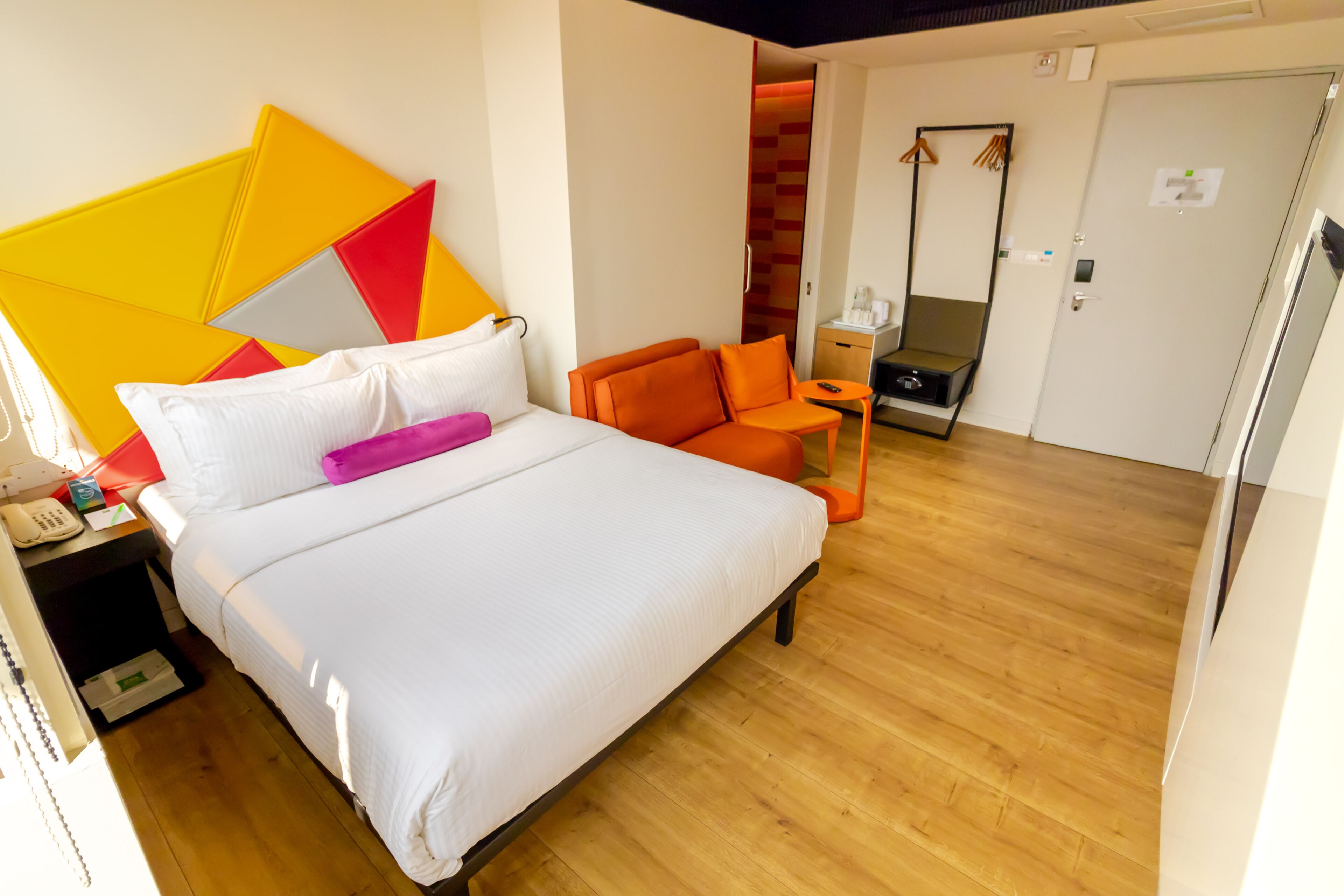 A hotel room with a bed, a nightstand, an orange chair, a small table, and a mirror.