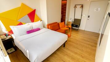 Una habitación de hotel con cama, mesita de noche, una silla naranja, una mesita pequeña y un espejo.