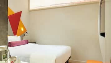 Habitación de hotel con cama, mesita de noche con una botella y una taza, almohada, un pequeño banco naranja y una lámpara de pared.