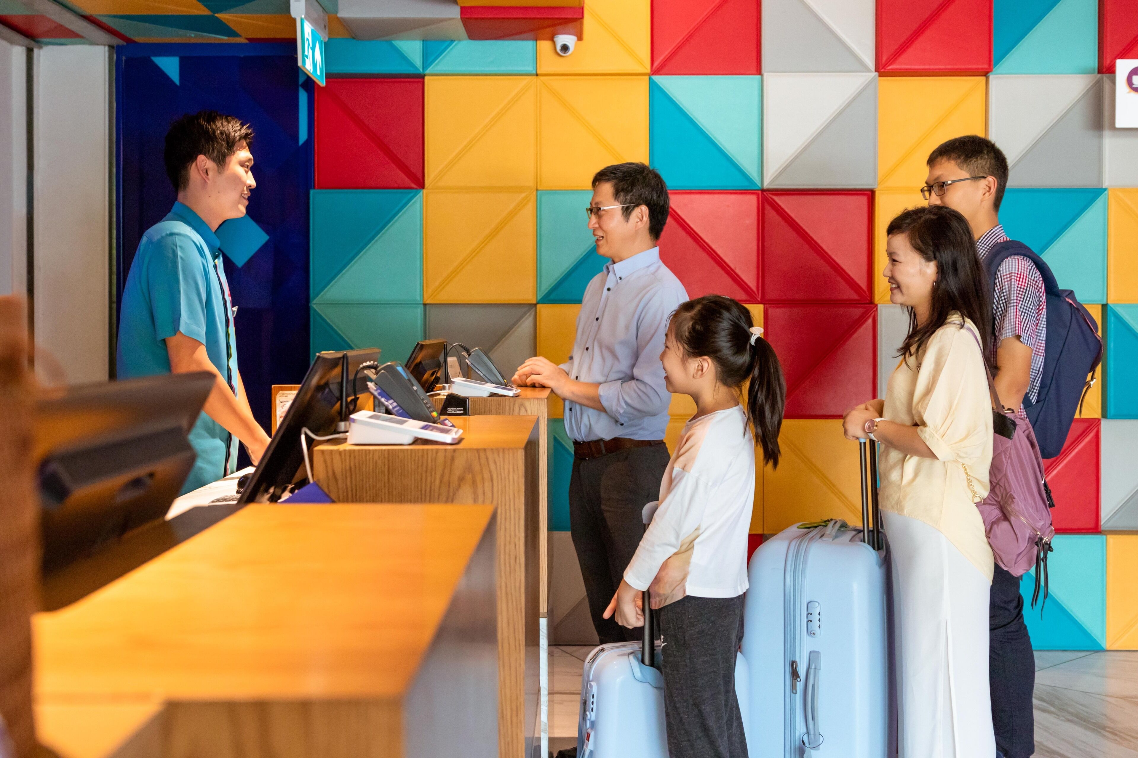 A reception desk with a wooden counter and a computer monitor. There are several people standing in line, some with luggage. The background features a colorful geometric wall design.