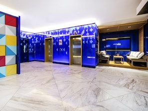 A modern lobby with a marble floor, elevators, a seating area with a TV, and a colorful geometric wall.