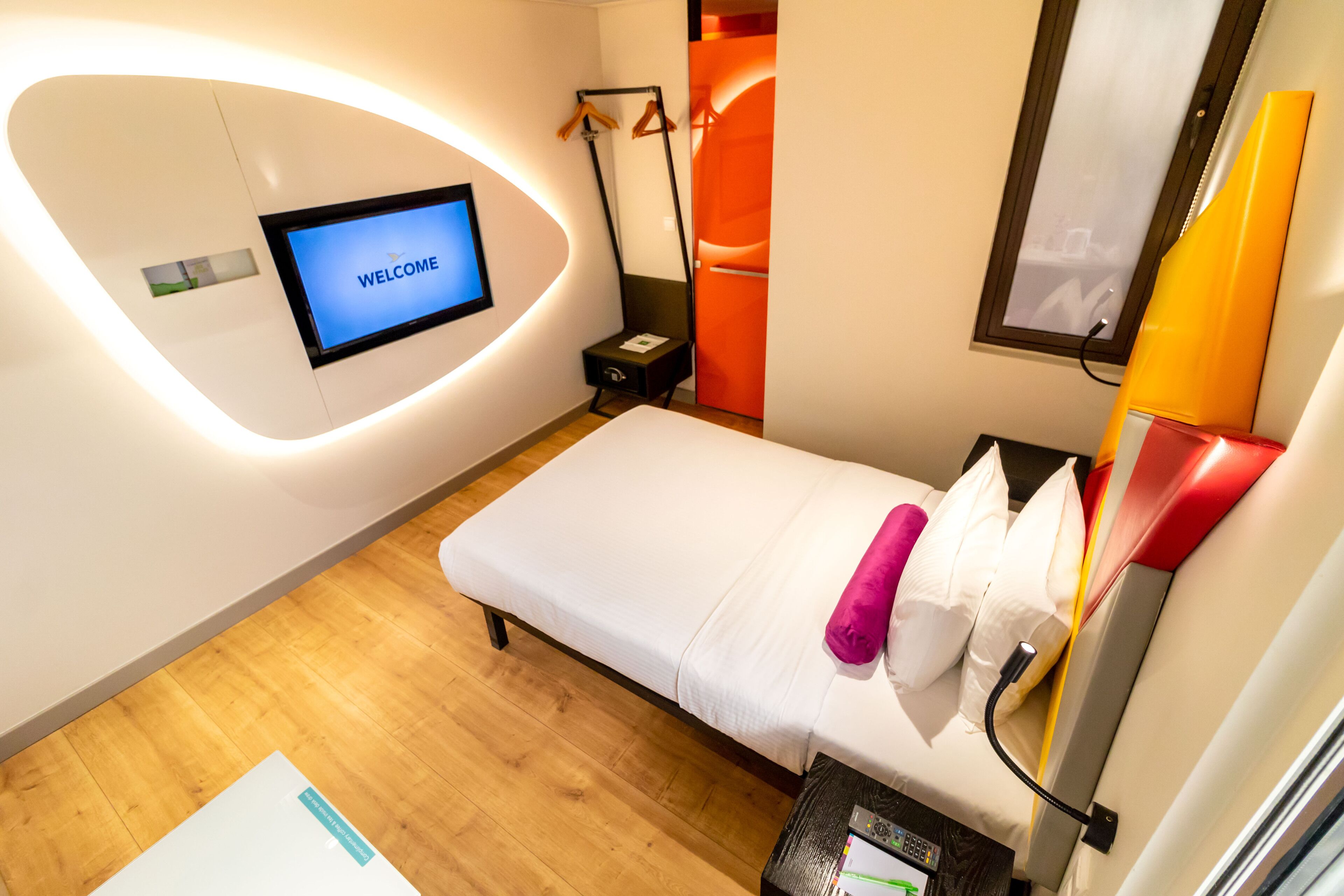 Ein modernes Hotelzimmer mit einem Bett, einem Fernseher, der "WELCOME" anzeigt, einem Nachttisch und einem an der Wand montierten Fernseher.