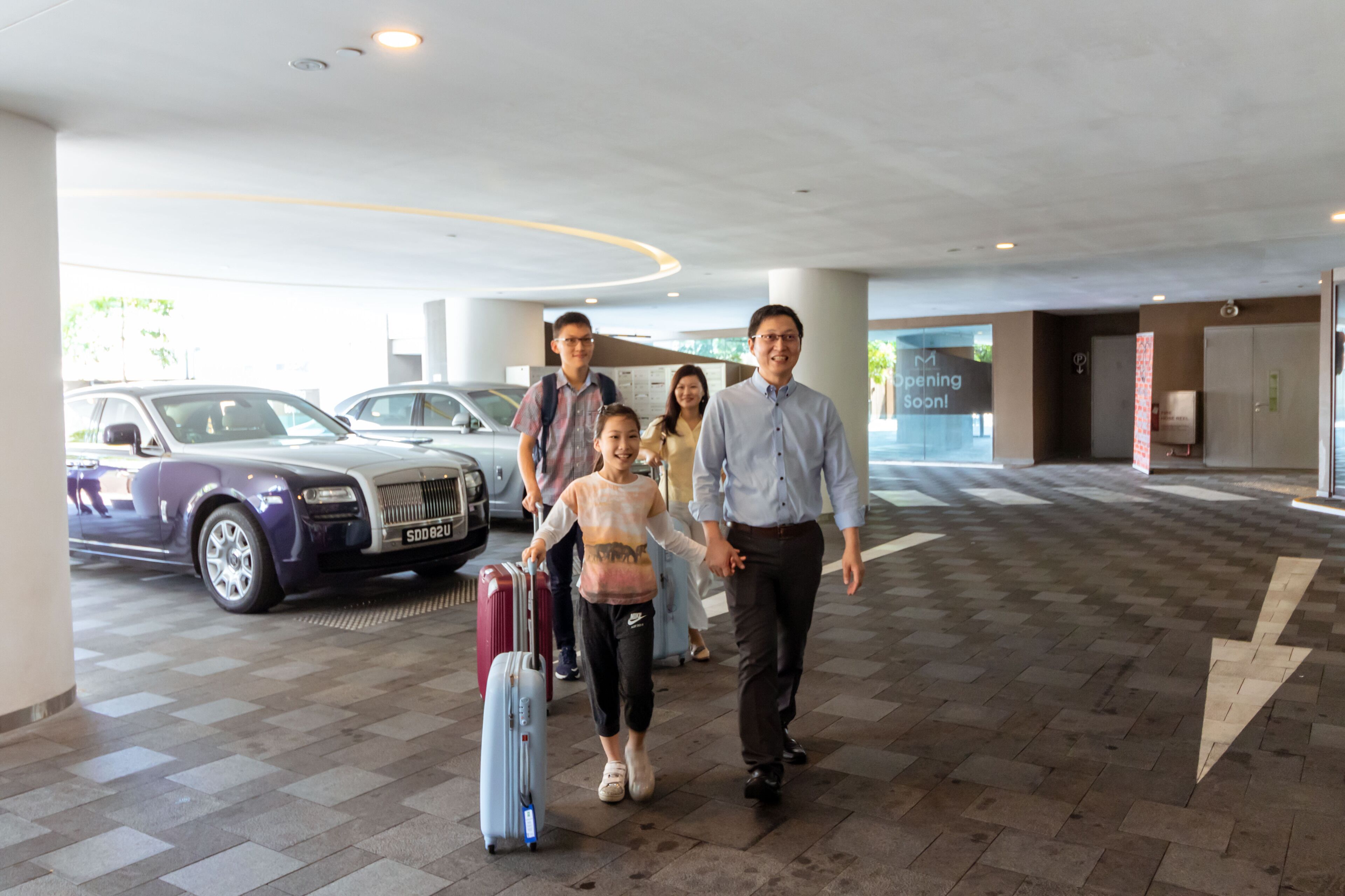 Een gezin loopt door de hotellobby, met een geparkeerde luxeauto en een bordje dat een nieuwe opening aankondigt.