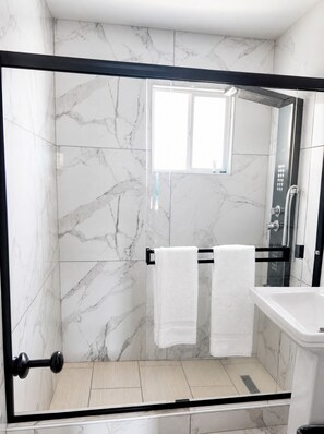 A modern bathroom with a glass-enclosed shower, marble walls, and a visible window. - Paradise Inn (Wildwood)