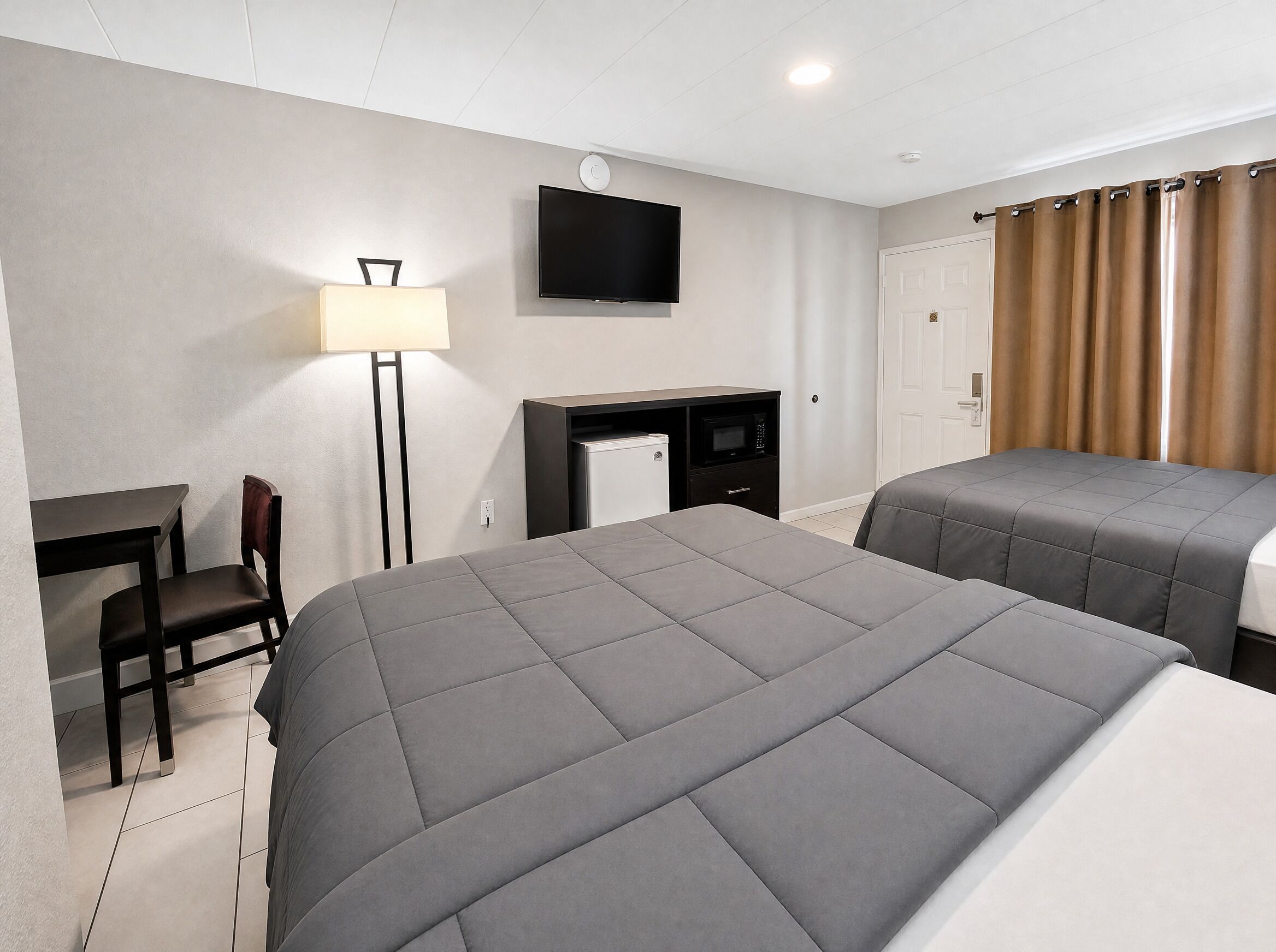 A hotel room with two beds, a desk, a chair, a TV, and a door with curtains.