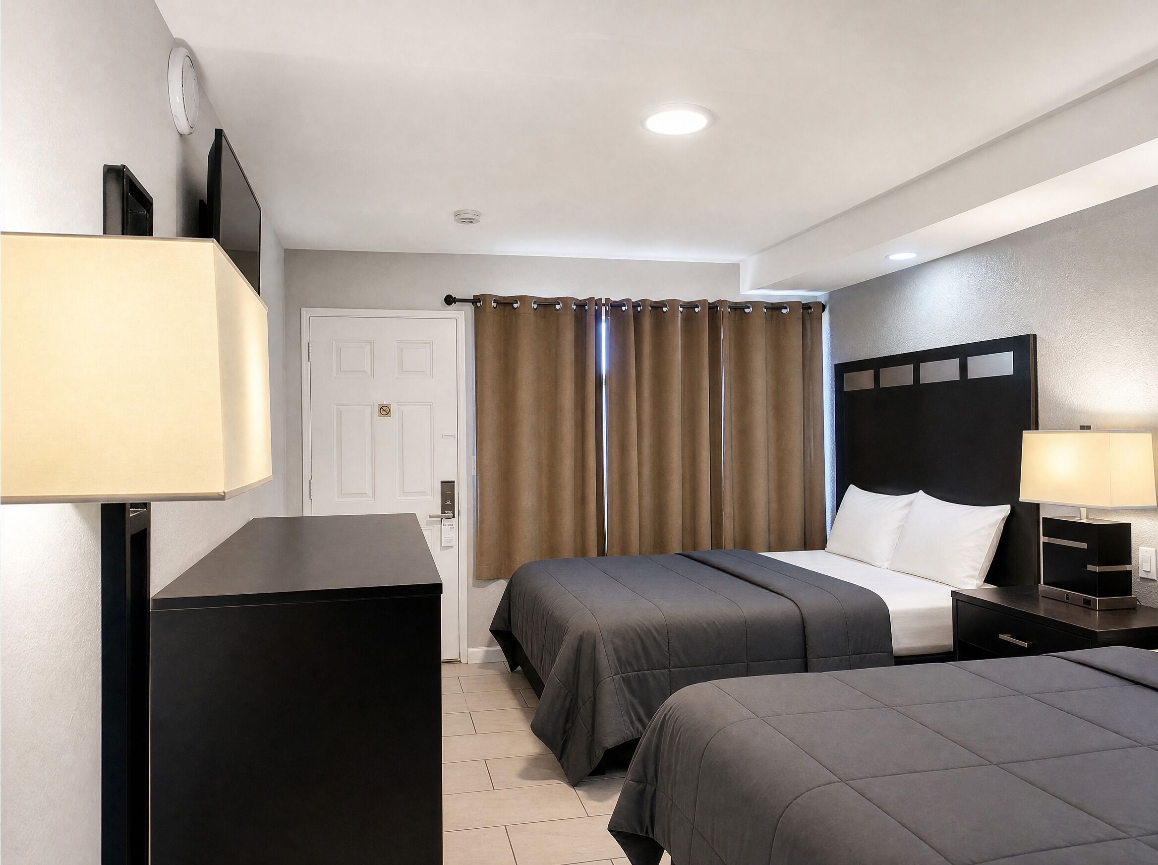 Habitación de hotel con dos camas, un escritorio, una lámpara, una puerta y una ventana con cortinas.