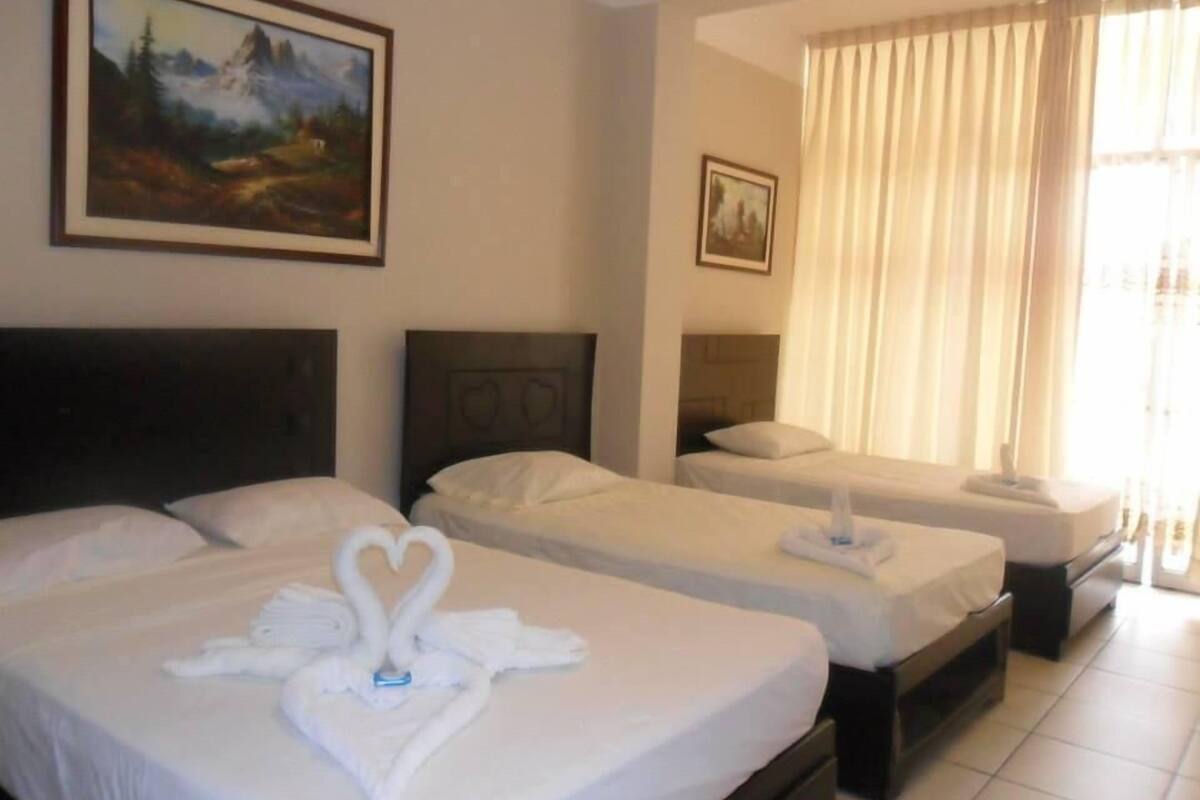 A hotel room with two beds, each with a heart-shaped towel arrangement, a painting on the wall, and a window with curtains.