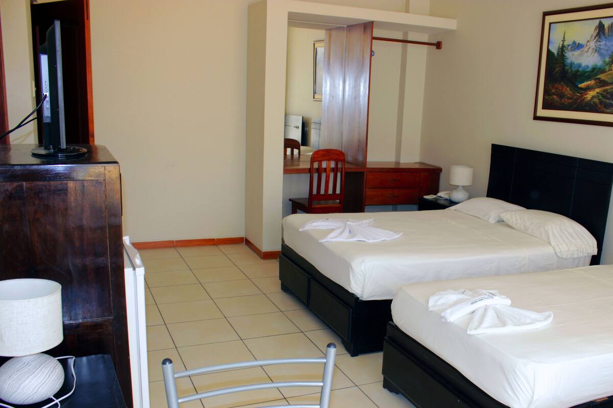 A hotel room with two beds, a desk, a chair, a TV, and a painting on the wall.