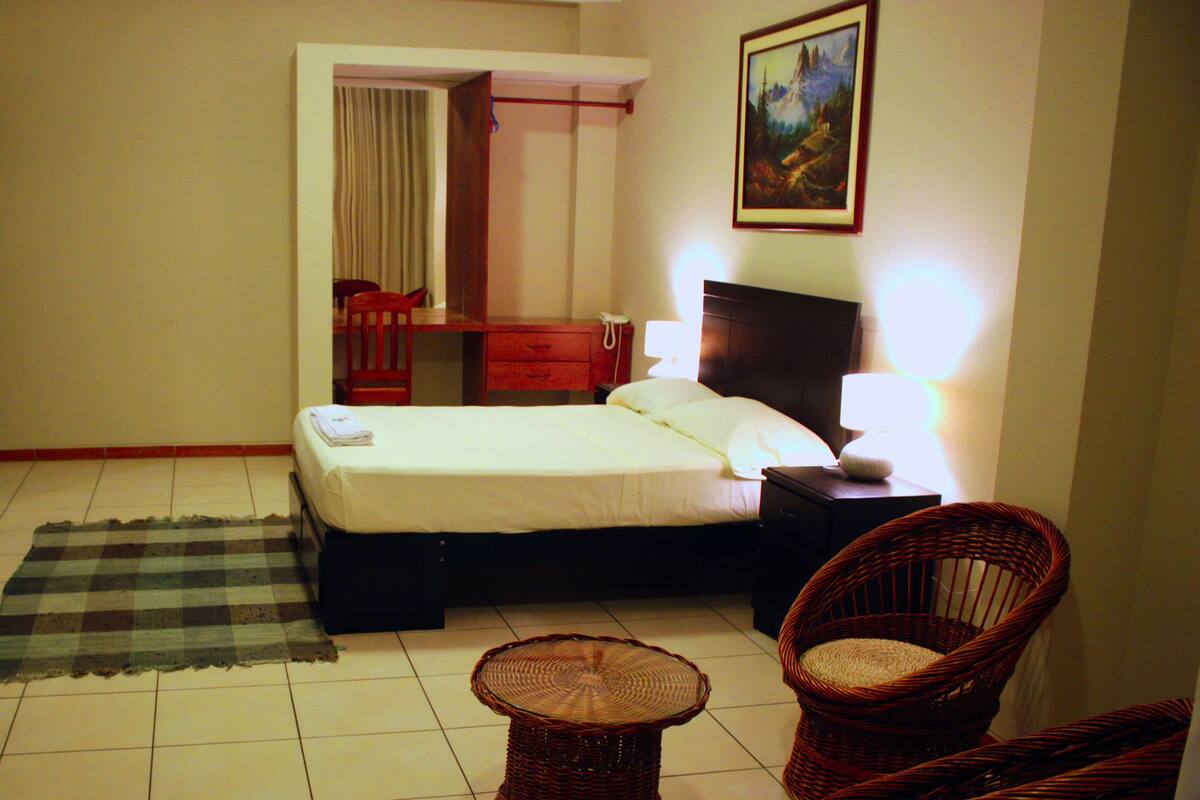 A hotel room with a bed, a desk, a chair, a TV, and a painting on the wall.