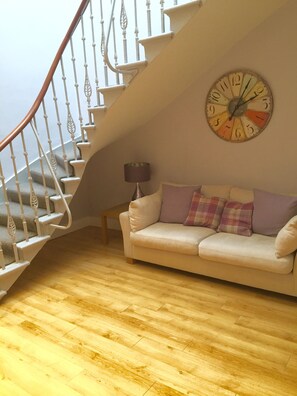 A staircase with white balusters, a round wall clock, and a beige sofa with plaid cushions.
