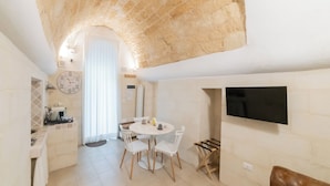 A modern kitchen with white cabinets, a round dining table with chairs, a flat-screen TV mounted on the wall, and a stone ceiling.