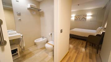Habitación de hotel compacta con ducha, inodoro y cama con mesita de noche y sillón.