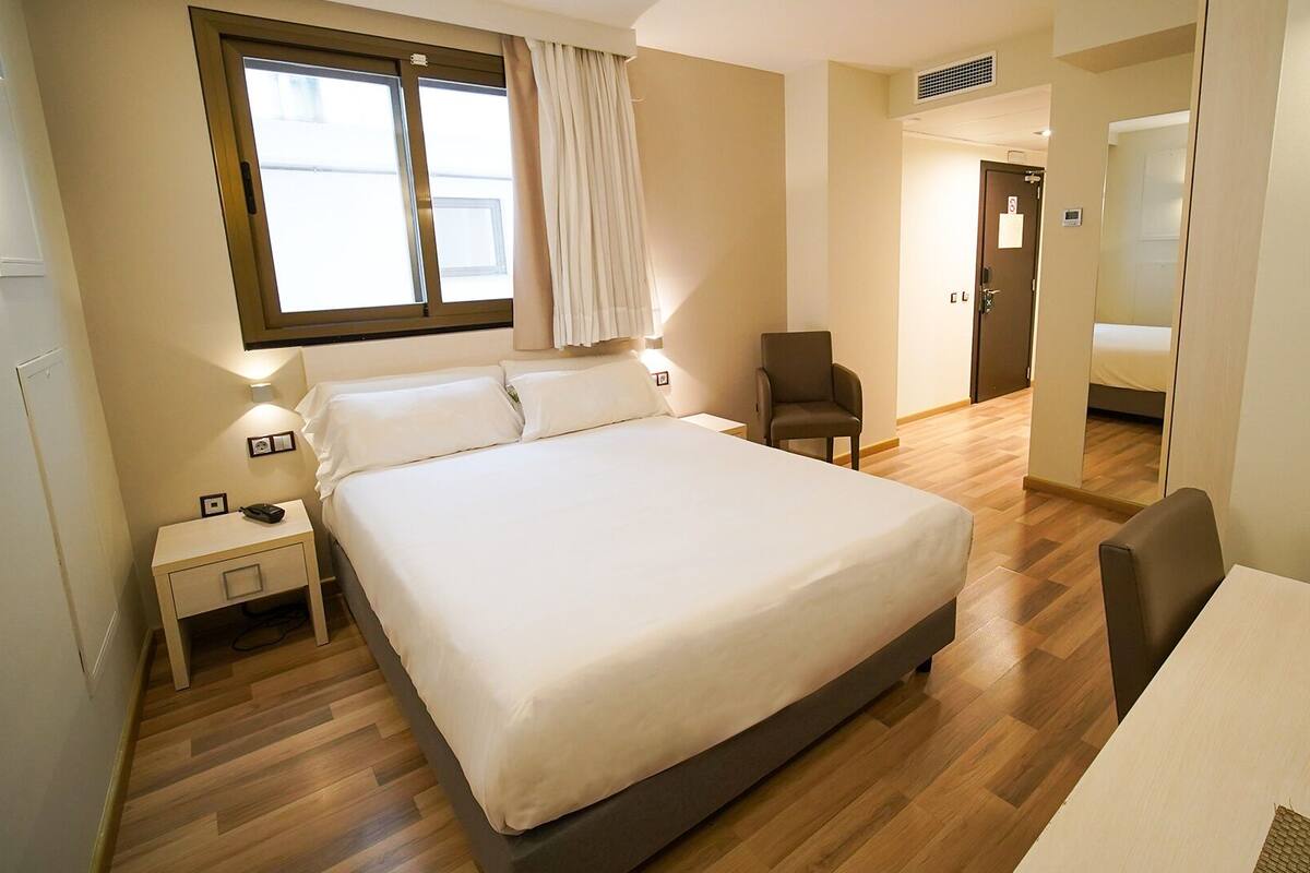A hotel room with a large bed, a chair, a desk, and a window with curtains.