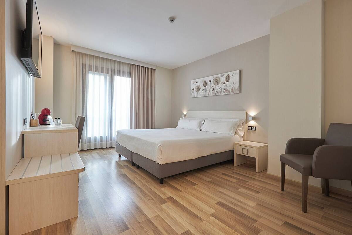 A hotel room with a bed, bedside tables, a desk, a chair, a TV, and a window with curtains.