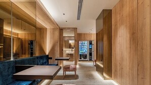 Un interior moderno con paneles de madera, una zona de estar con un banco y un reposapiés, una máquina expendedora y un ascensor visible.