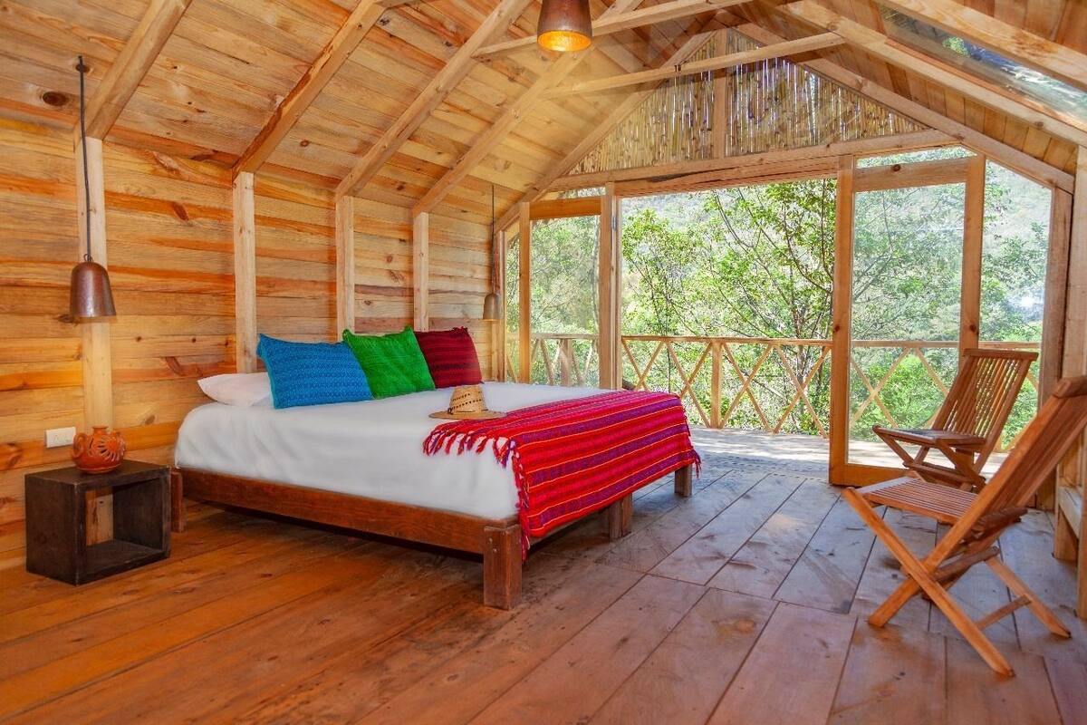 Un dormitorio de cabaña de madera con cama, almohadas, una mesita de noche, una silla y un ventanal grande con vistas a follaje.