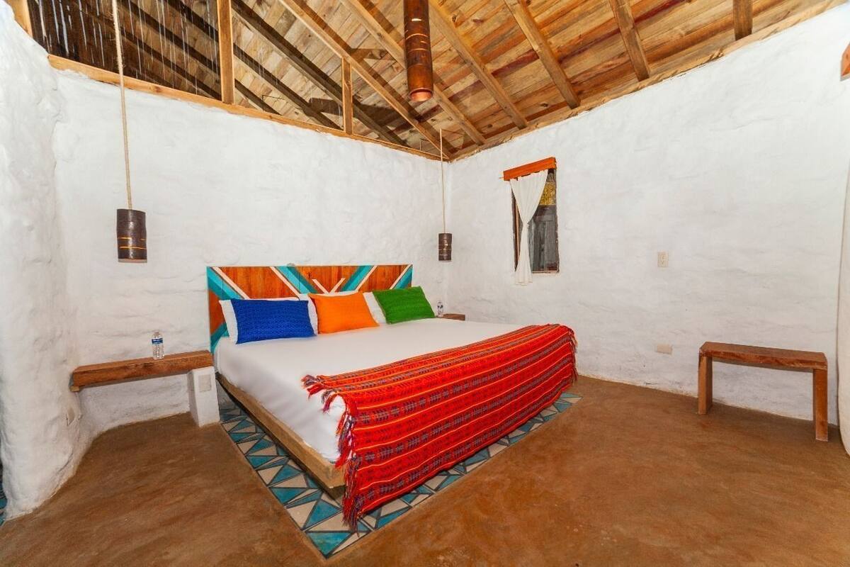 Una habitación con cama, vigas de madera, paredes blancas y una lámpara colgante.