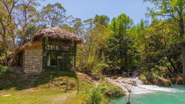 A rustic stone house with a thatched roof, situated by a clear turquoise river, surrounded by lush greenery. - Canto del Agua Ecolodge (Tzimol)