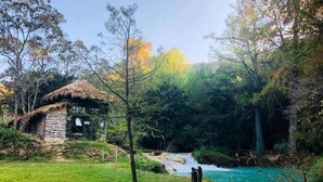 A rustic stone hut with a thatched roof, surrounded by lush greenery and a flowing river. - Canto del Agua Ecolodge (Tzimol)
