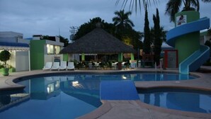A swimming pool with lounge chairs and a thatched-roof structure, surrounded by palm trees and buildings. - Hotel York (Guamuchil)