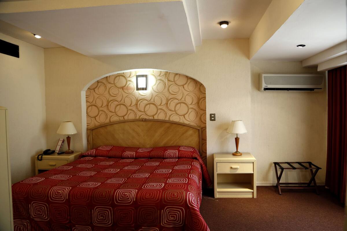 A hotel room with a patterned headboard, a bed with a red and white bedspread, bedside tables with lamps, a small shelf, and an air conditioning unit.