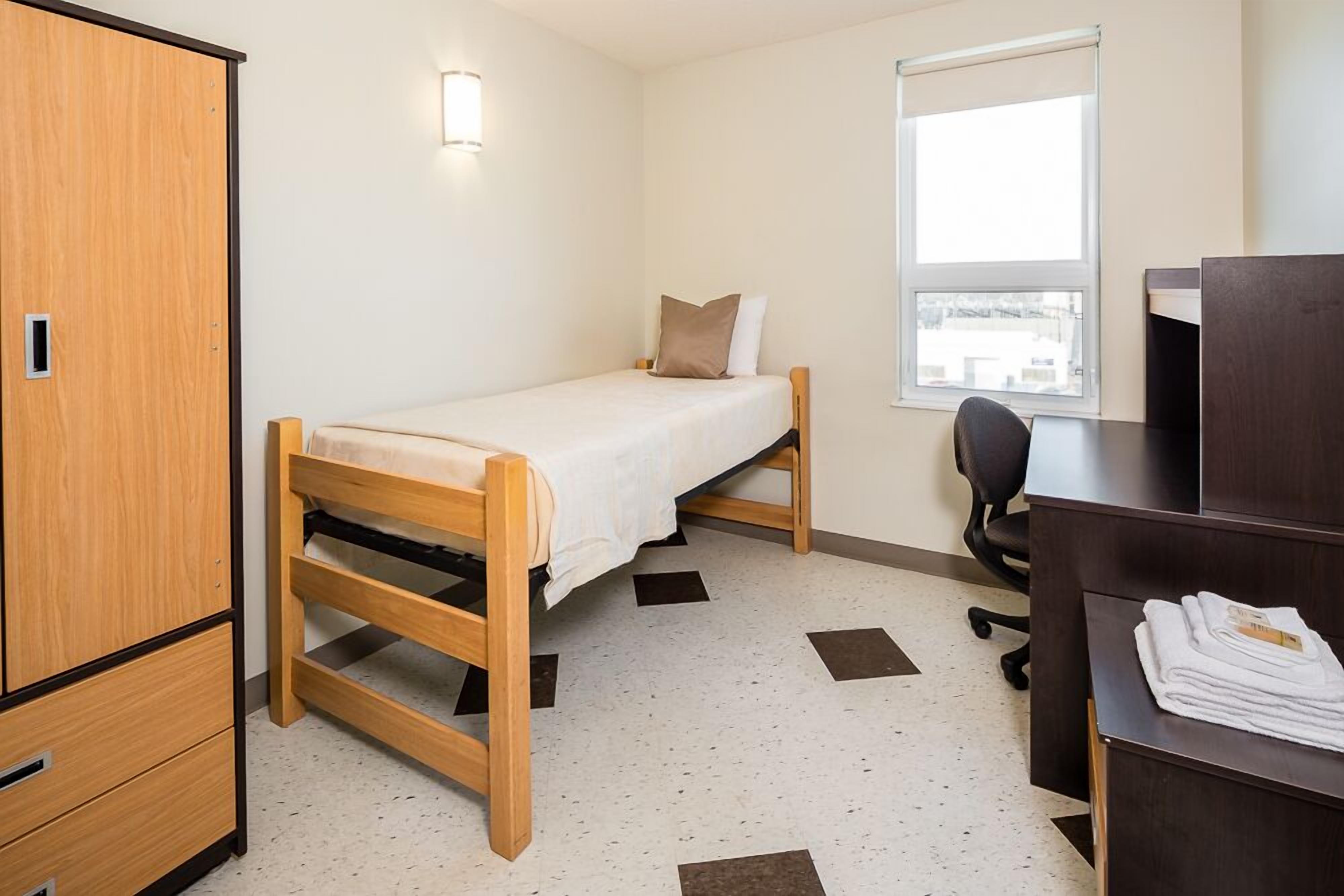 A single-bed room with a wooden bed, a desk, a chair, a wardrobe, and a window with a view.