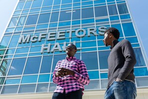 Deux personnes se tiennent devant un bâtiment doté d’une grande façade en verre, sur laquelle on peut lire l’inscription « MCFEETORS HALL ».