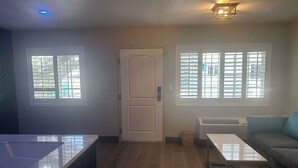A room with a wooden floor, a white door, a window with blinds, a white table, and a sofa. - Super 8 by Wyndham Oceanside Downtown (Oceanside)