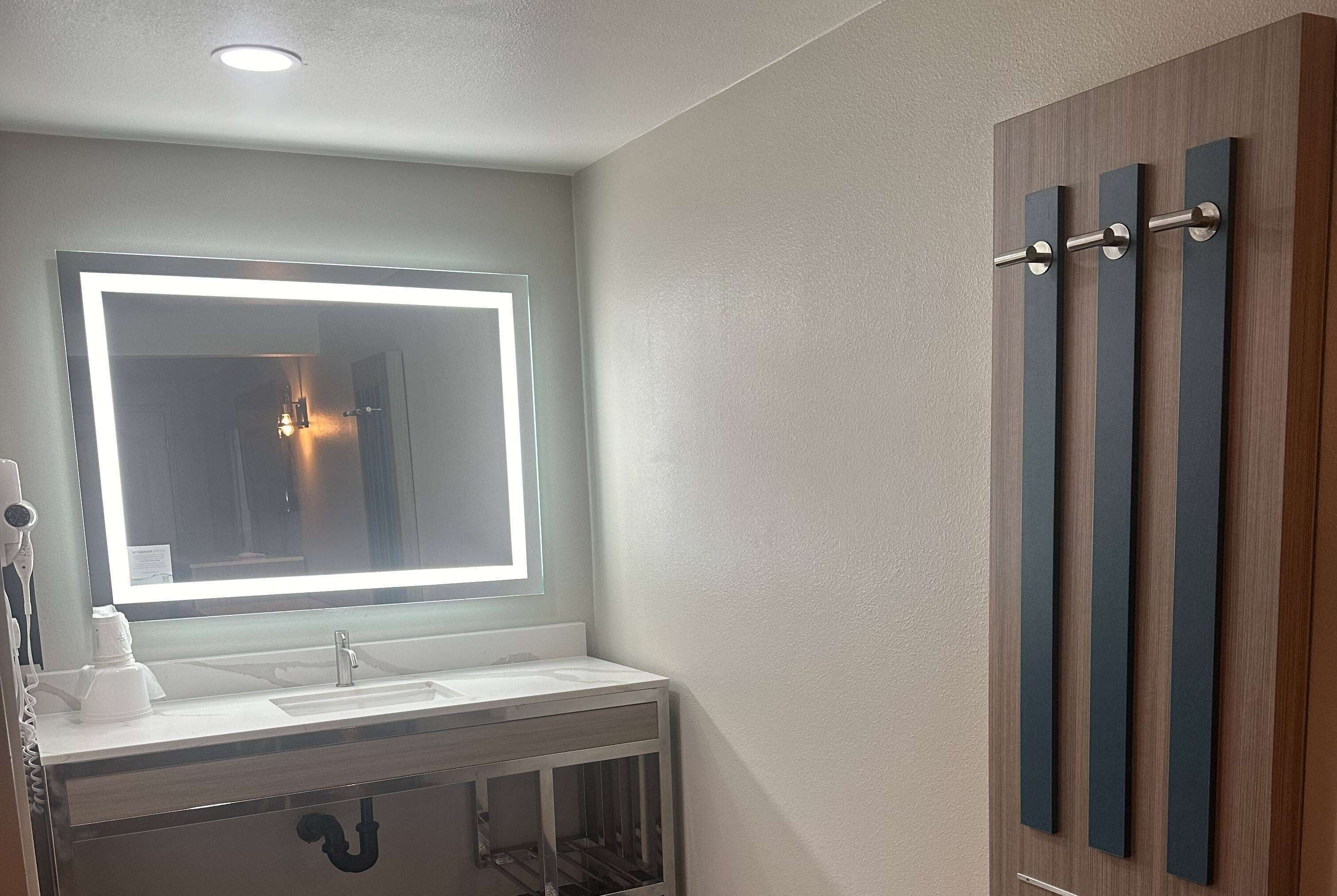 Uma casa de banho com uma toucador moderno, um espelho grande com iluminação integrada, uma pia e um armário com porta de vidro.