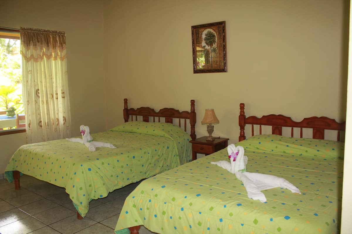 Deux lits jumeaux avec des draps verts et des serviettes blanches disposés en forme de cygnes, une tête de lit en bois, une petite table de chevet avec une lampe et un tableau encadré au mur.