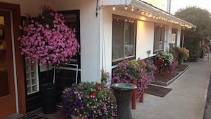 Un edificio con fachada blanca, grandes ventanales y un porche decorado con flores rosadas colgantes y plantas en macetas.