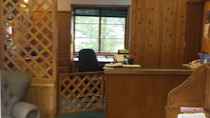 Una habitación acogedora con un escritorio de madera, una silla y un sofá. Se observa una ventana con persianas, una alfombra verde y una partición de madera.