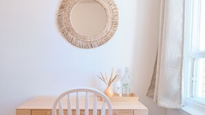 Un espacio de trabajo pequeño y ordenado con un escritorio de madera, una silla blanca, un espejo redondo tejido y un radiador.