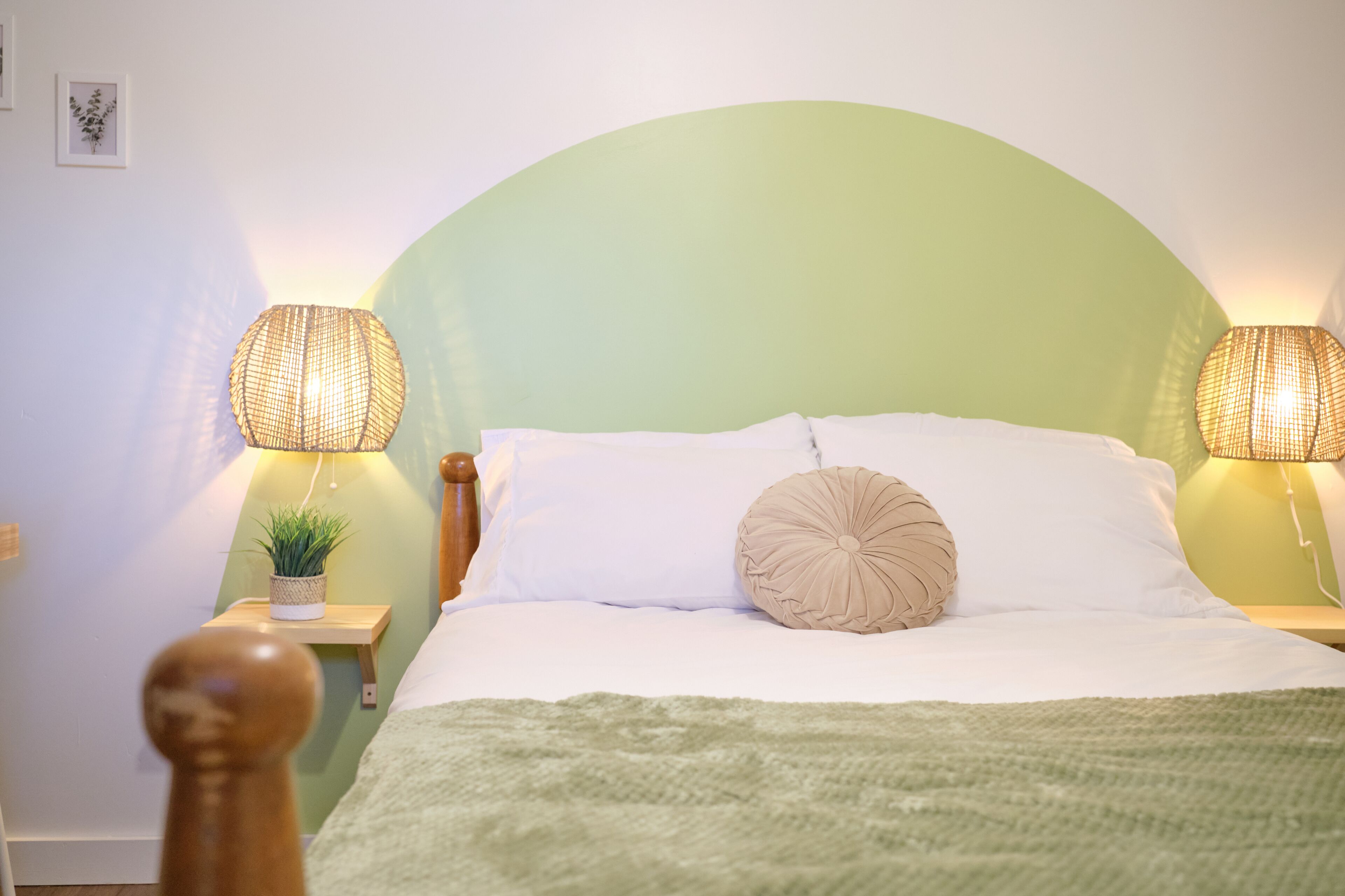 A neatly made bed with white linens and a green throw blanket, flanked by two bedside lamps with woven shades.