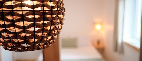 Een detail van een geweven lamp met een warme gloed, een bed met een groene sprei en een raam met daglicht.