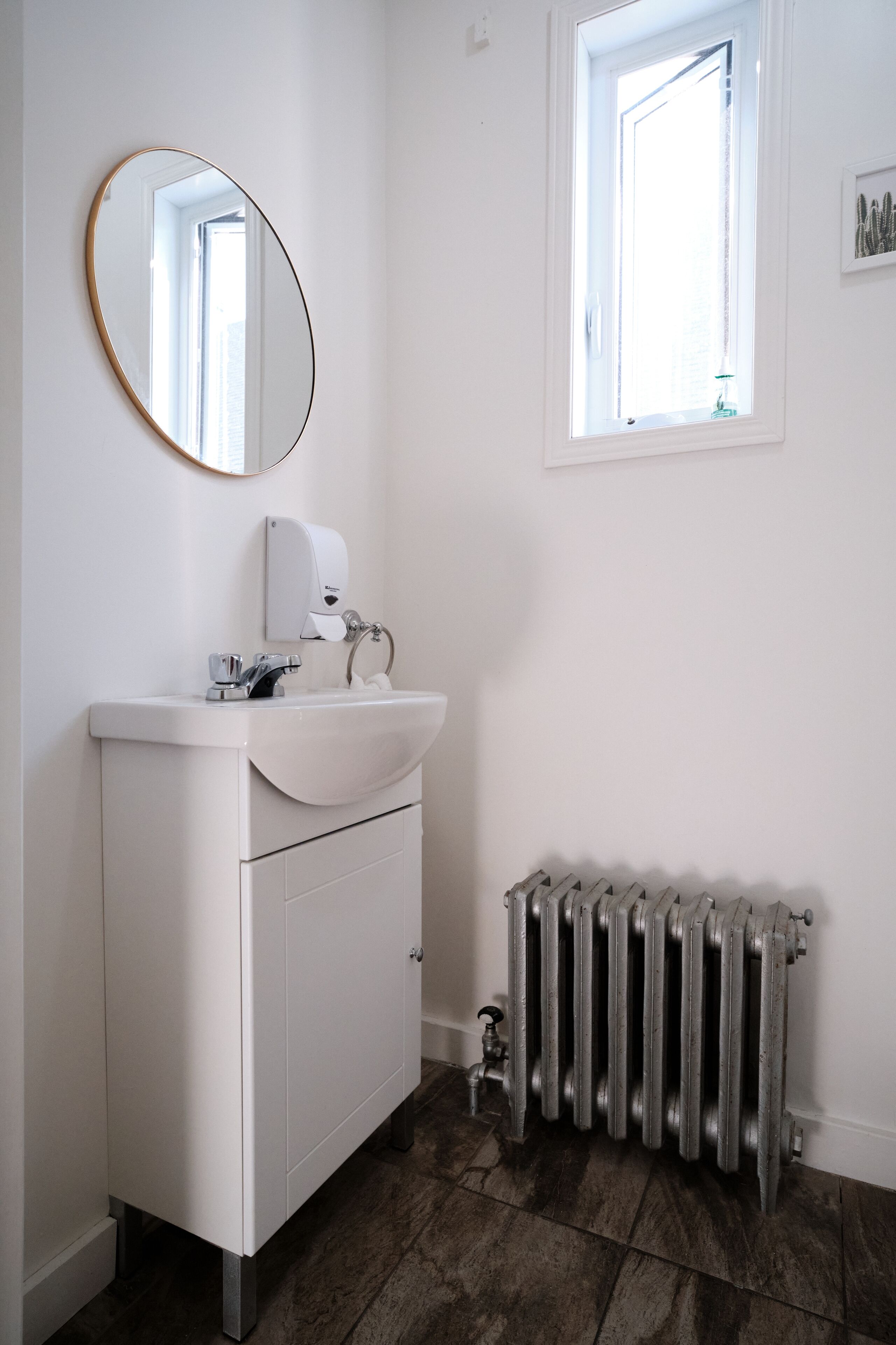 A bathroom with a white vanity, a round mirror, a window, and a radiator.