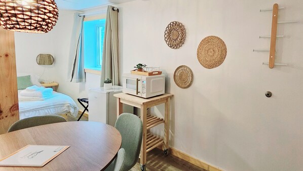 A compact hotel room with a bed, a small table with a chair, a microwave on a wooden cart, and decorative wall hangings.