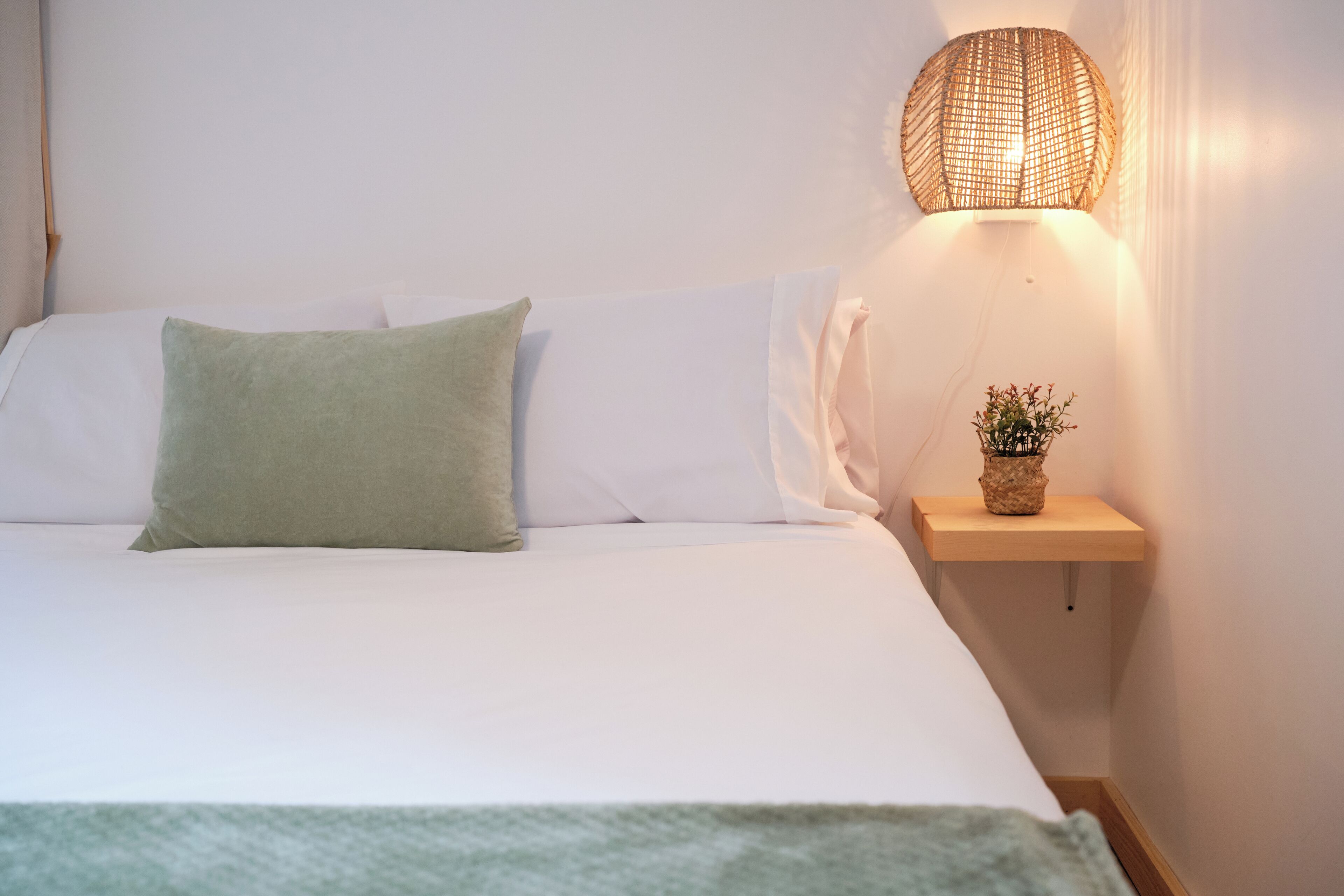 A neatly made bed with white and green pillows, a small wooden nightstand with a potted plant, and a woven wall lamp.