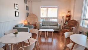 Una sala de estar acogedora con un sofá gris, sillas blancas, un sillón redondo de mimbre, una mesita blanca y una lámpara de pie.
