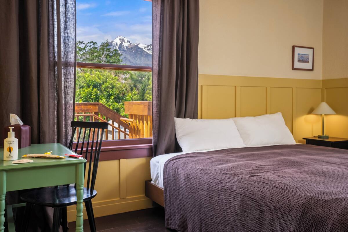 A hotel room with a bed, a table with a chair, a window with a view of mountains, and a framed picture on the wall.