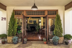 Entrada de un hotel con puerta de madera, plantas en macetas, chimenea de piedra y una escalera.