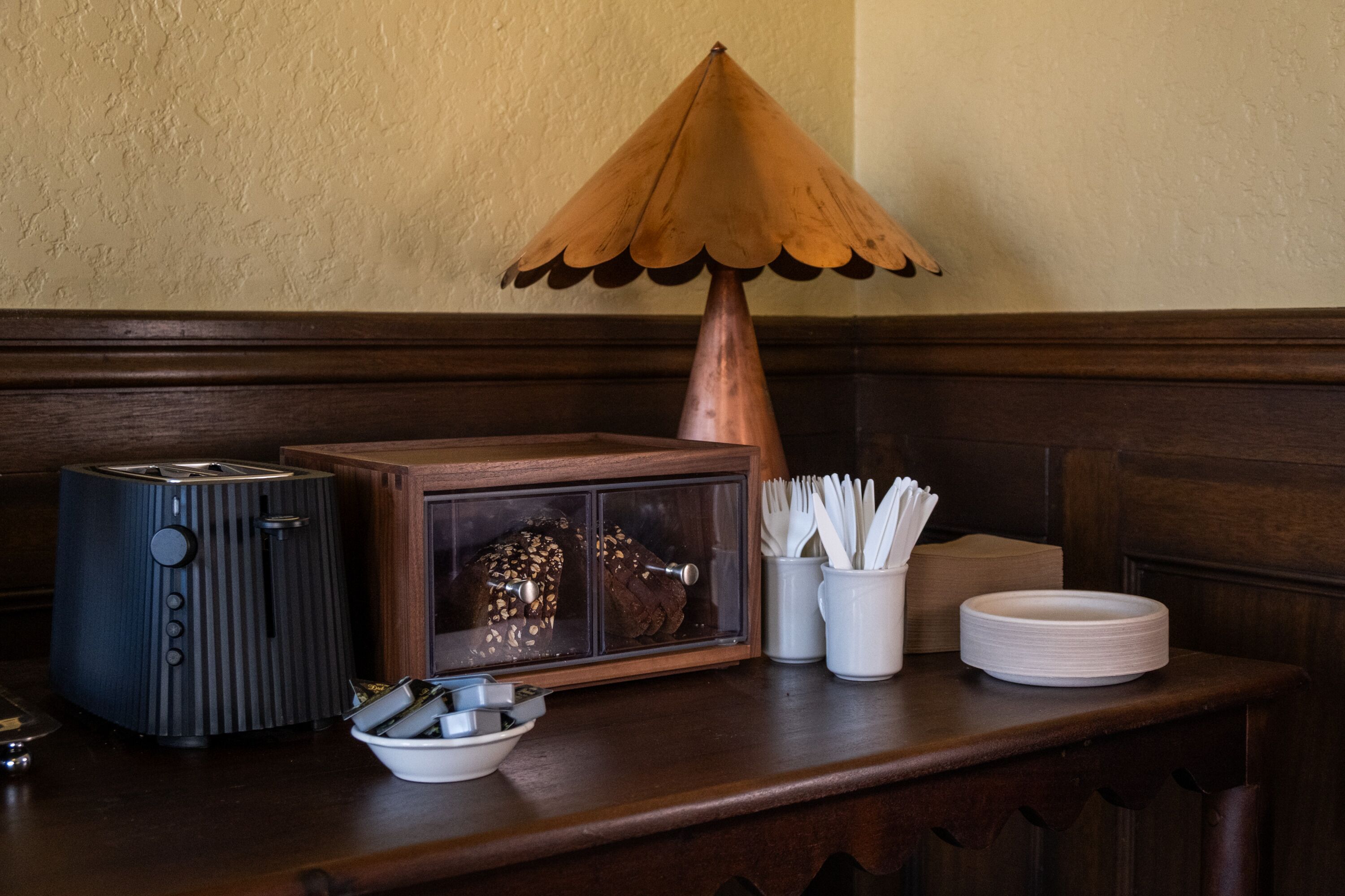 A wooden table with a toaster, a bread box, a bowl, a plate, and a cup with utensils.